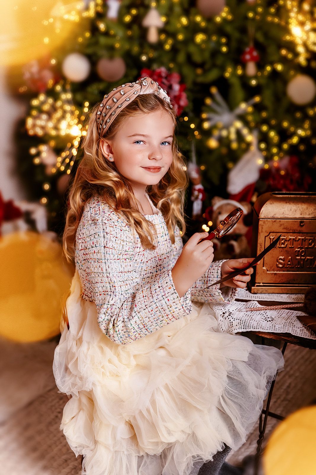 Young girl in white tulle skirt poses by decorated Christmas tree with vintage mailbox, holding magnifying glass.