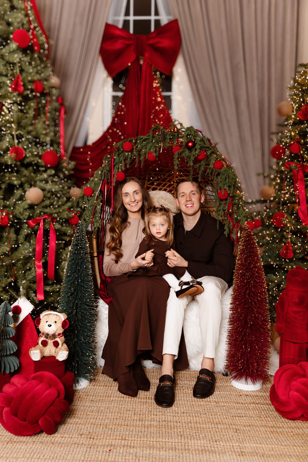 Family of three posing together on a chair surrounded by festive Christmas trees and red holiday decorations.