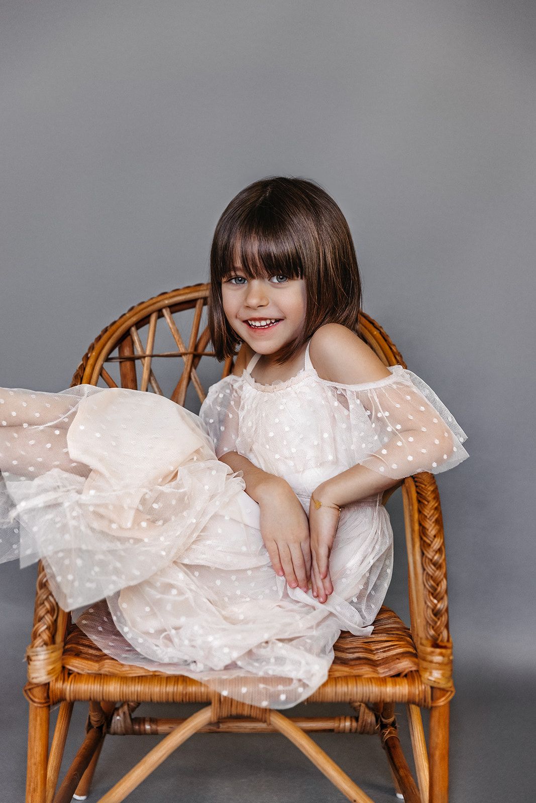 Young girl in white polka dot tulle dress sitting in wicker chair, smiling at camera against gray background.