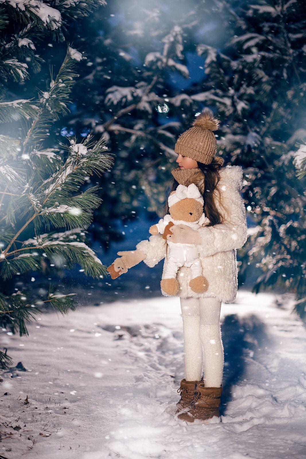 A young girl in white winter outfit holds a teddy bear among snow-covered pine trees at night.