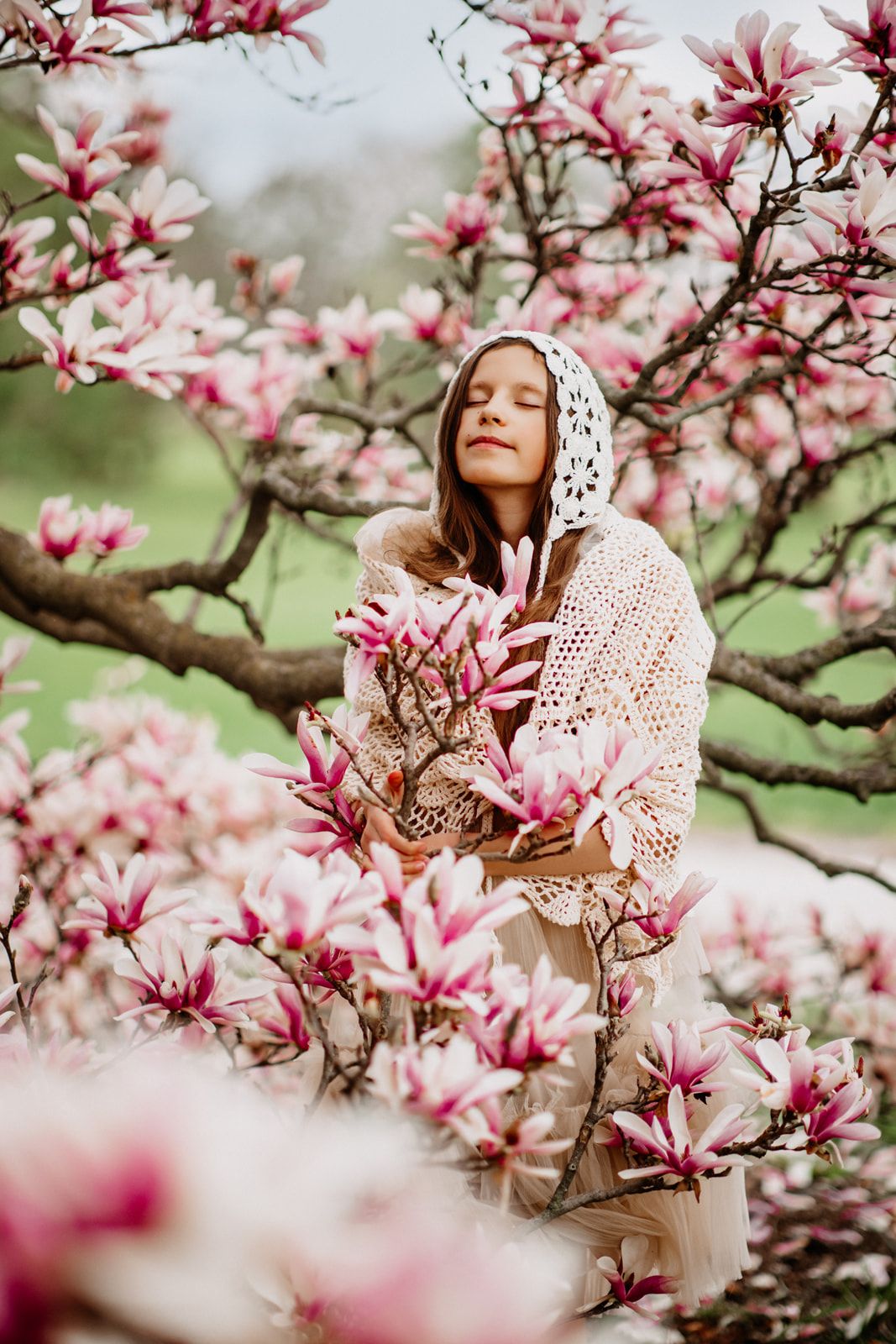Young girl with eyes closed embraces pink magnolia flowers wearing white crochet wrap in spring garden.