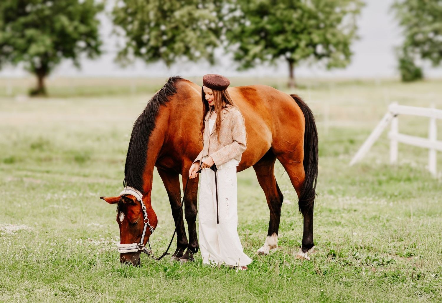 Woman in white dress and hat stands beside a bay horse grazing in a lush green field.