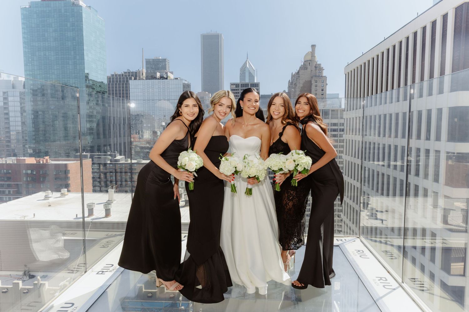 Bride in strapless white gown poses with four bridesmaids in black dresses on rooftop with Chicago skyline.