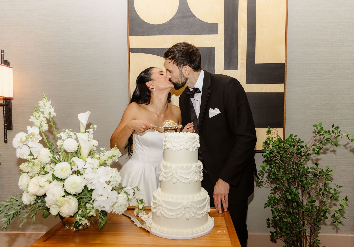 Bride and groom sharing a kiss while cutting their elegant white tiered wedding cake at their reception.