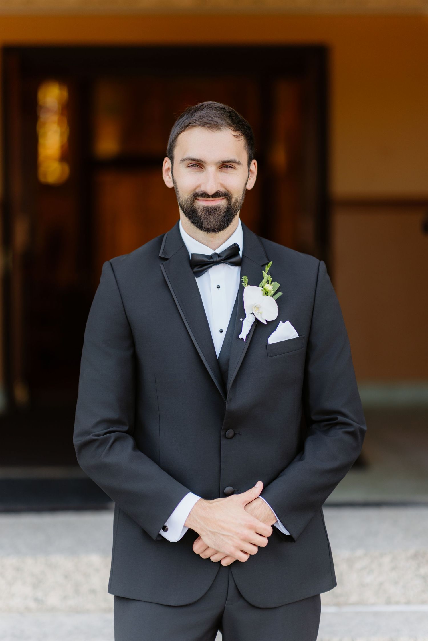 Handsome bearded groom in black tuxedo with white boutonniere smiling outside church entrance.
