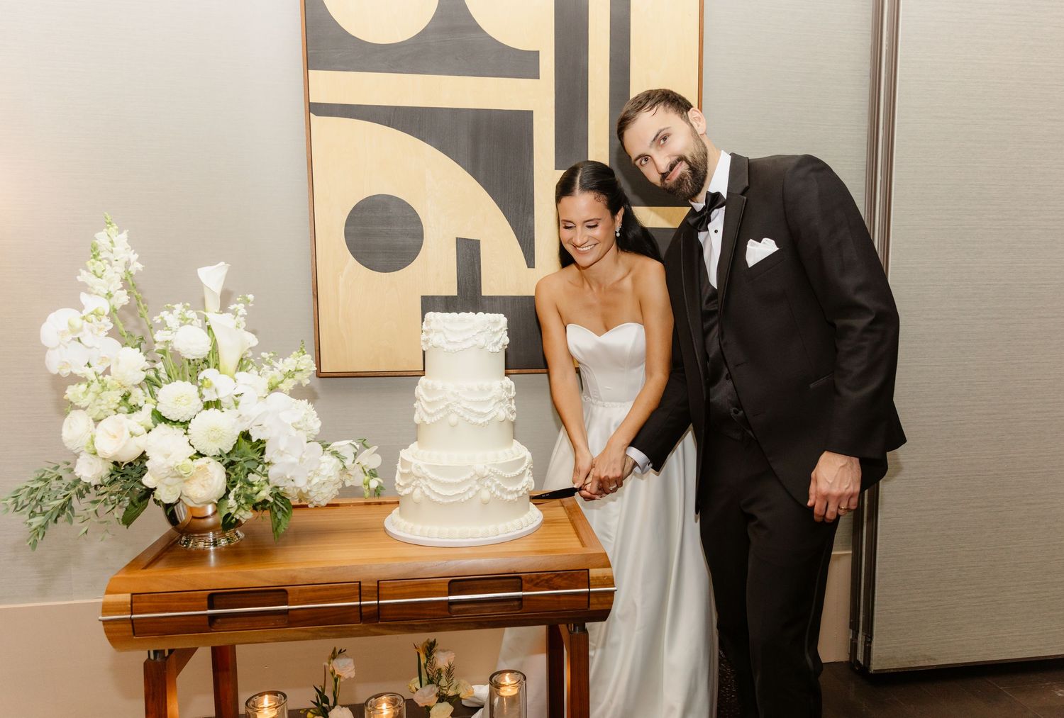 Bride and groom cut elegant white tiered wedding cake adorned with lace details beside white floral arrangement.