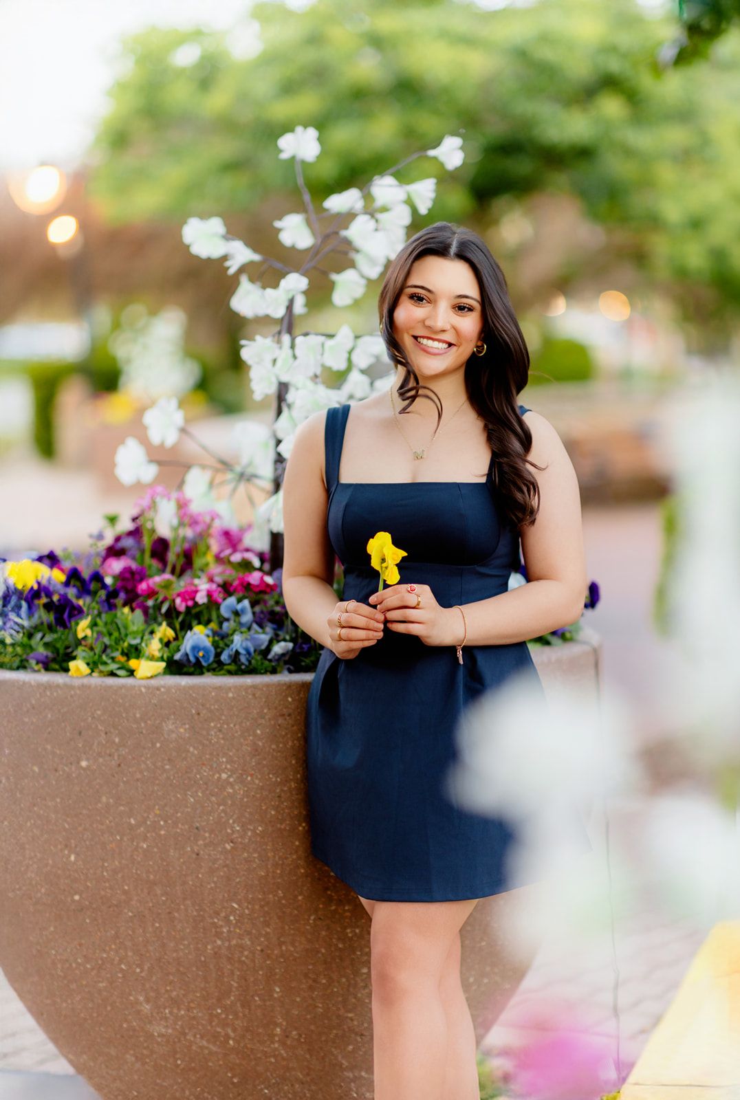 Young woman in navy dress smiling while holding a yellow flower near colorful blooms.