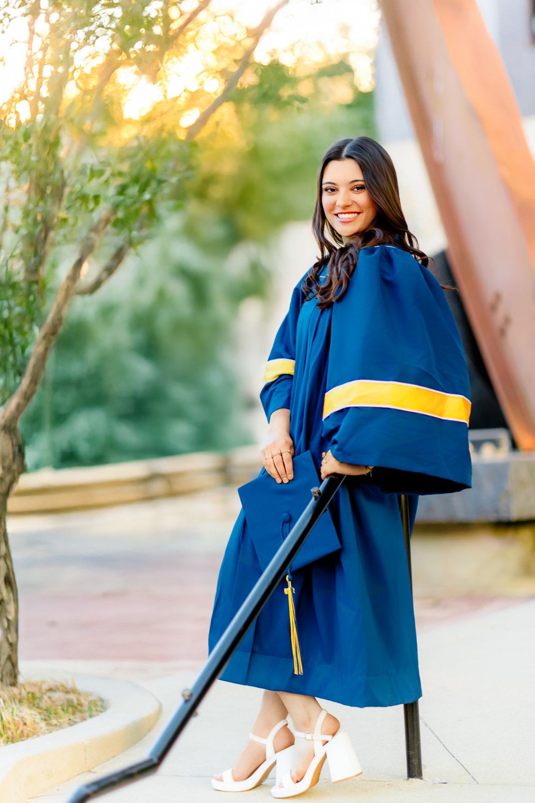 Smiling graduate in navy blue and gold graduation gown posing outdoors at golden hour.