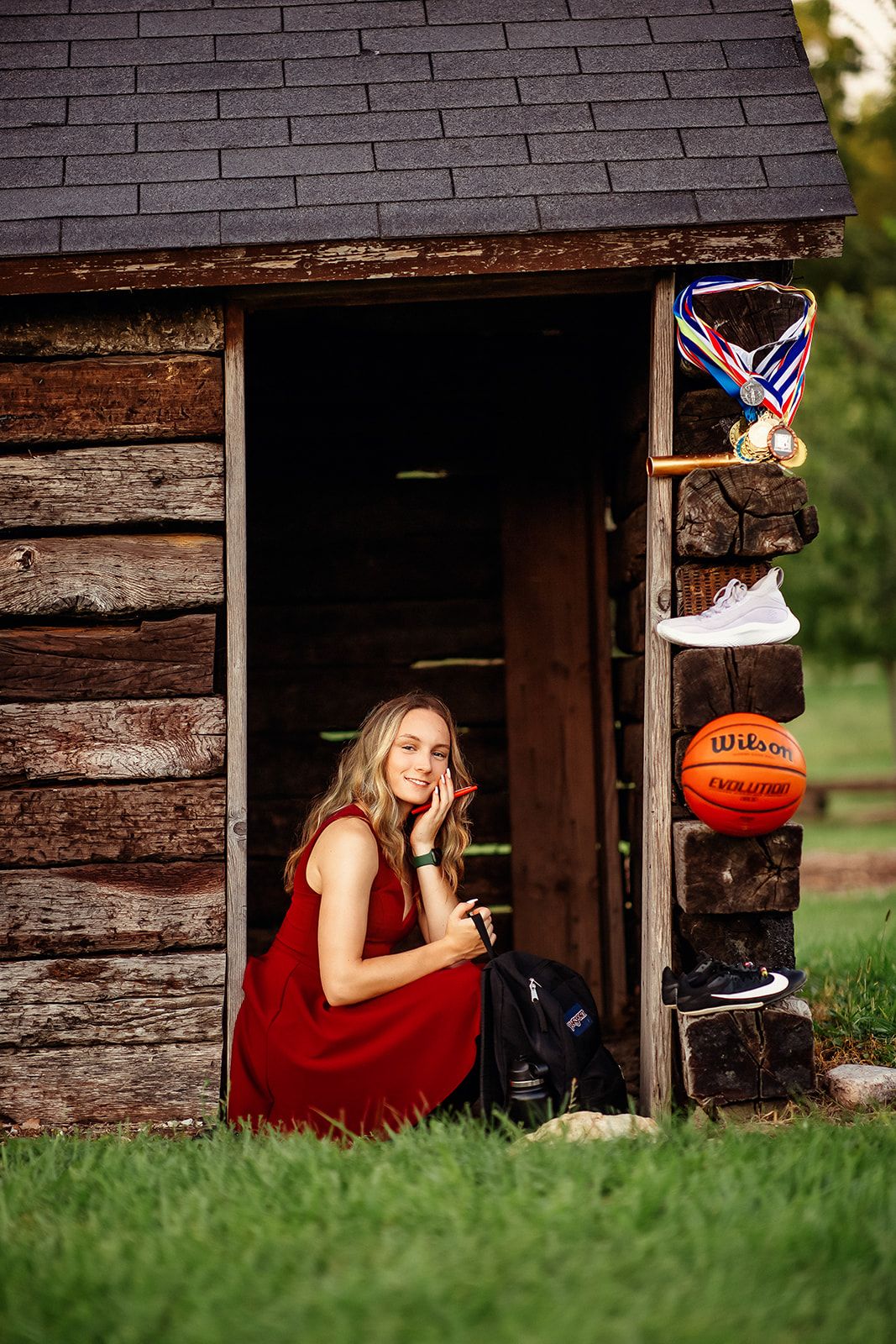 Teen girl in red dress sitting in log cabin doorway with basketball and medals displayed.