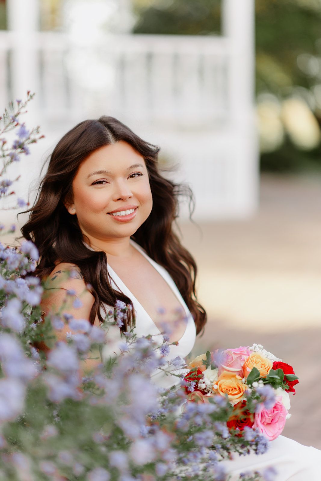 Smiling bride with dark wavy hair holding colorful bouquet of roses surrounded by purple wildflowers near white gazebo.