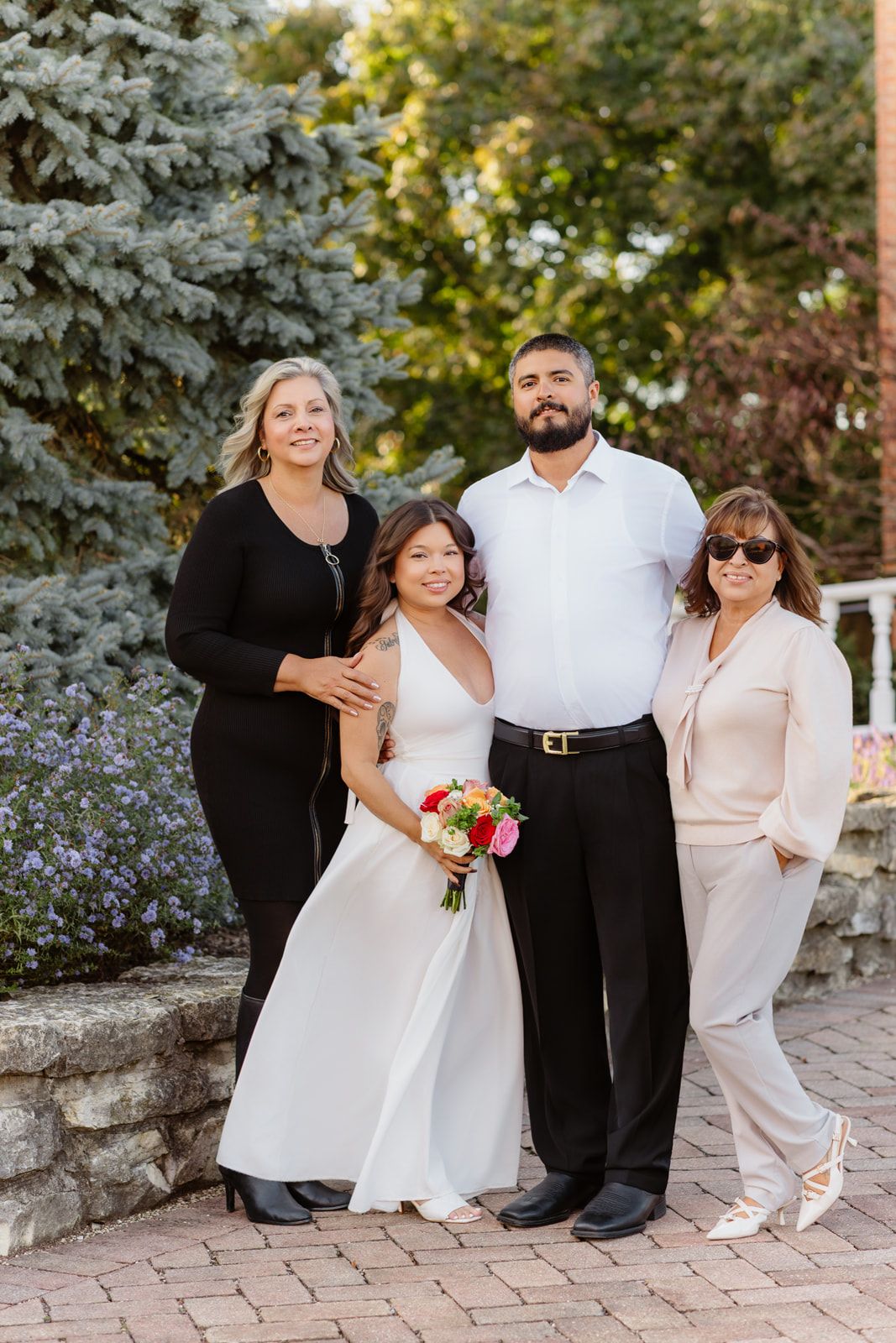 Bride and groom pose with two women in formal attire in an outdoor garden setting.