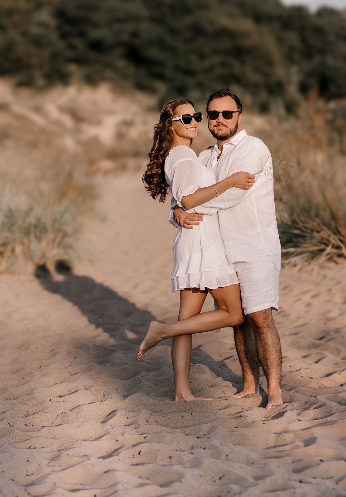 Couple in white outfits embracing on a sandy beach path with sunglasses during golden hour.