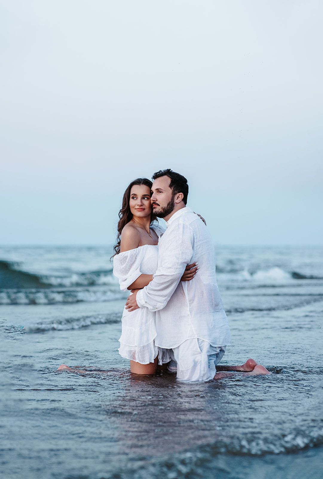 Romantic couple in white outfits embracing in ocean waves on a beach at dusk.
