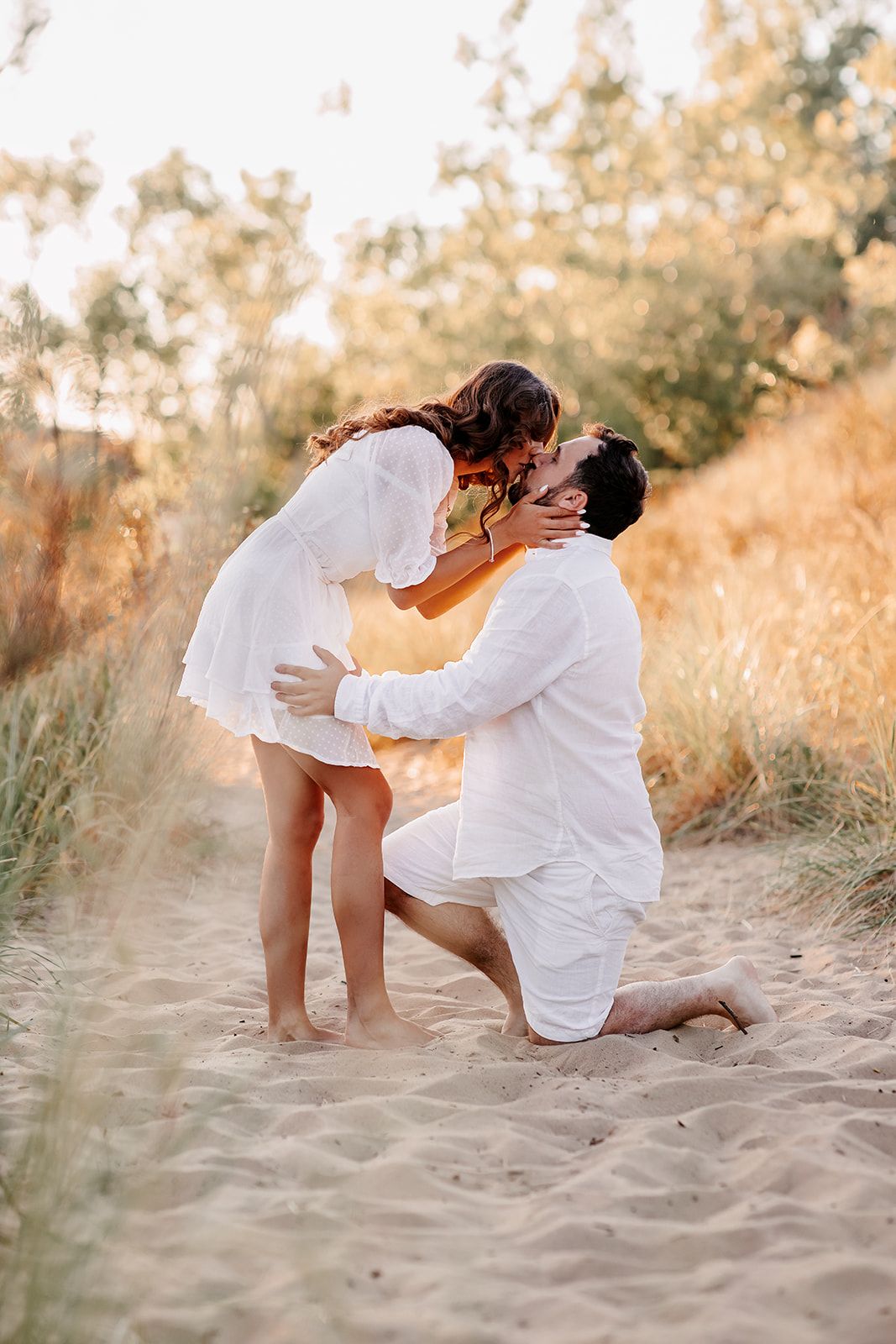 Man on one knee proposing to woman on sandy beach path with golden grass backdrop.