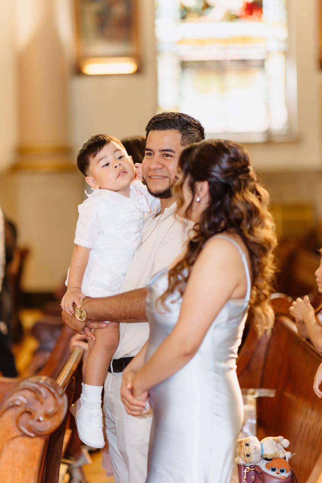 Smiling father holds young child in white outfit beside mother in church during baptism celebration.