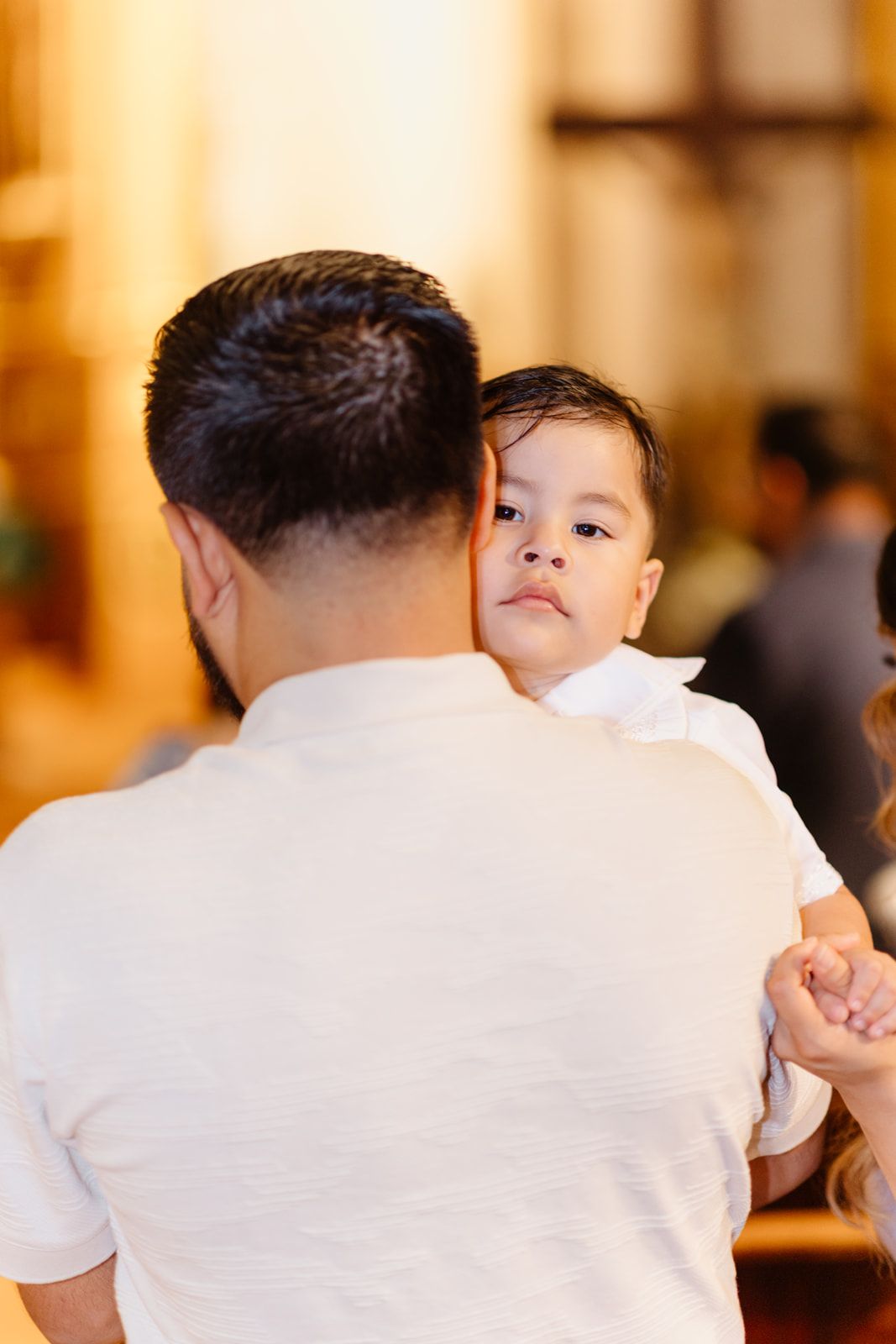 A man in a white shirt holds a young toddler in a church setting, the child resting their head on his shoulder.