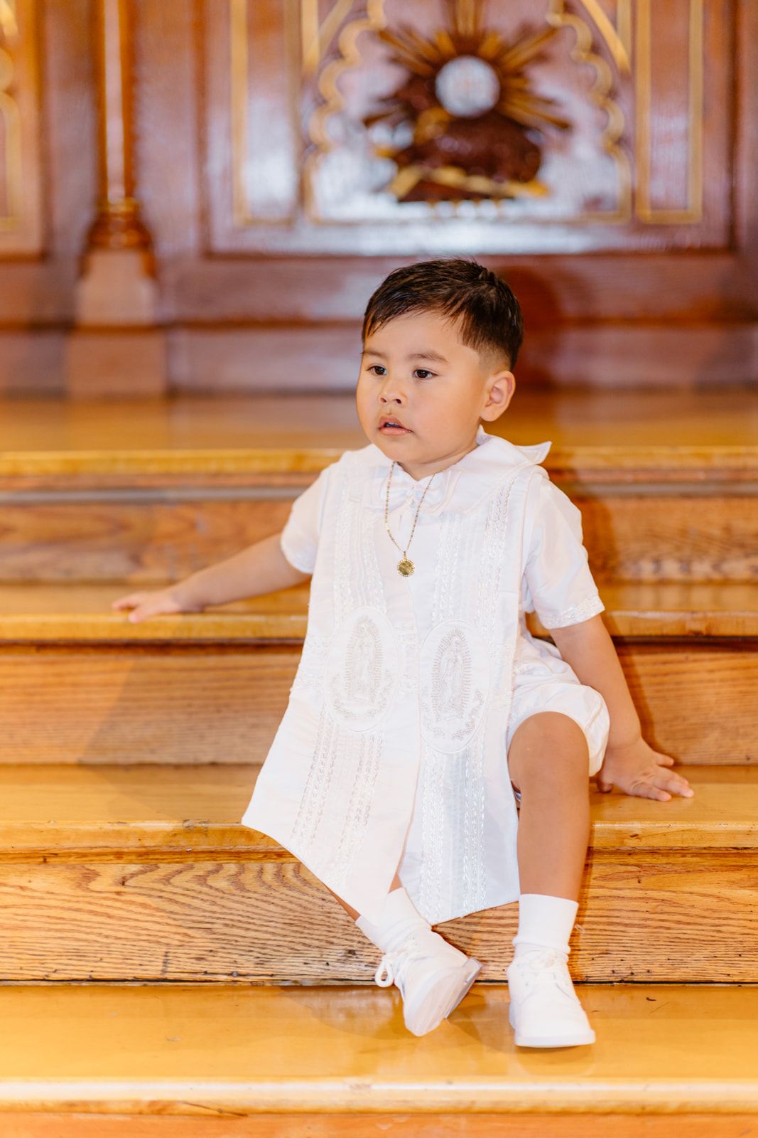 Toddler boy in white embroidered Filipino barong sits on wooden church altar steps wearing a necklace.