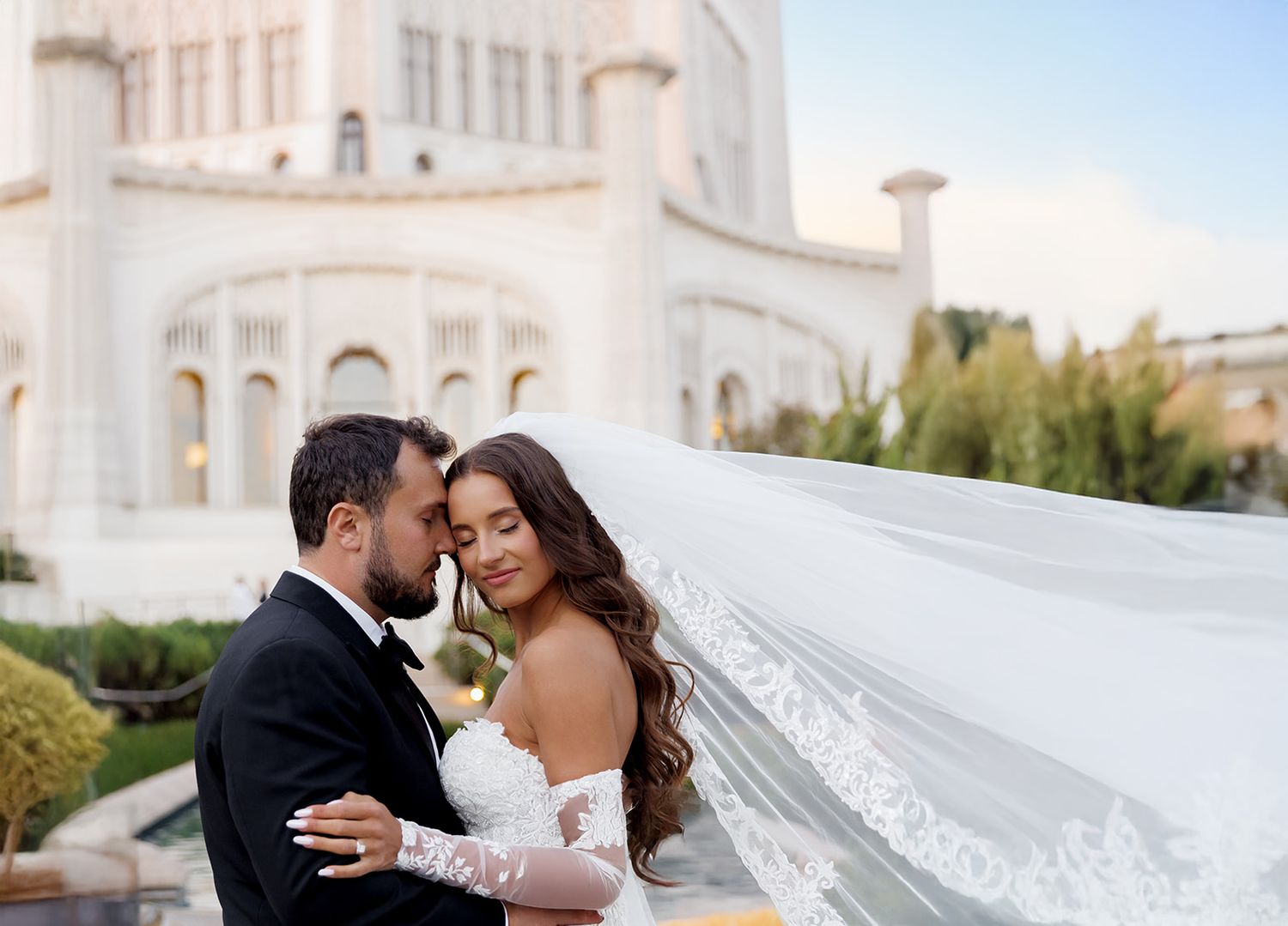 Romantic bride and groom touch foreheads tenderly as her veil flows near a grand white temple.