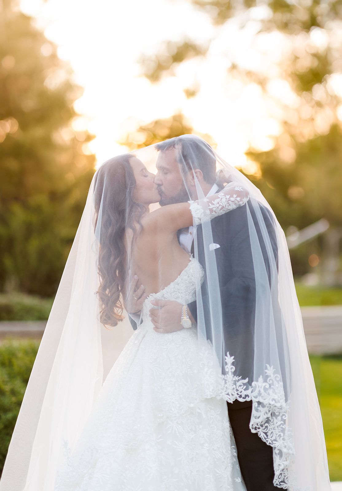 Bride and groom kissing under a lace-trimmed veil at golden hour, with lush greenery in the background.
