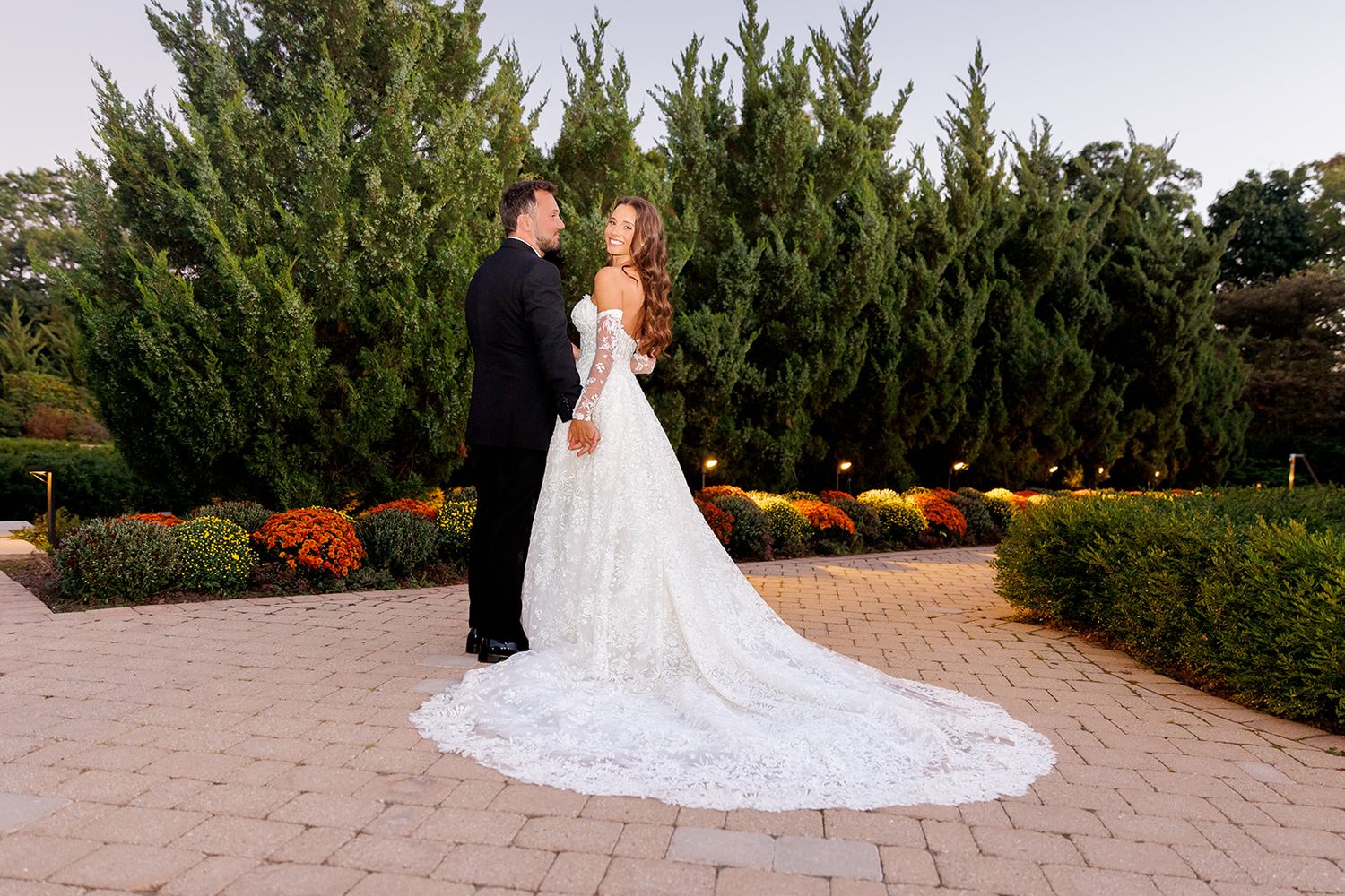 Bride in white gown and groom in tuxedo share a romantic moment on a garden path surrounded by tall evergreens.