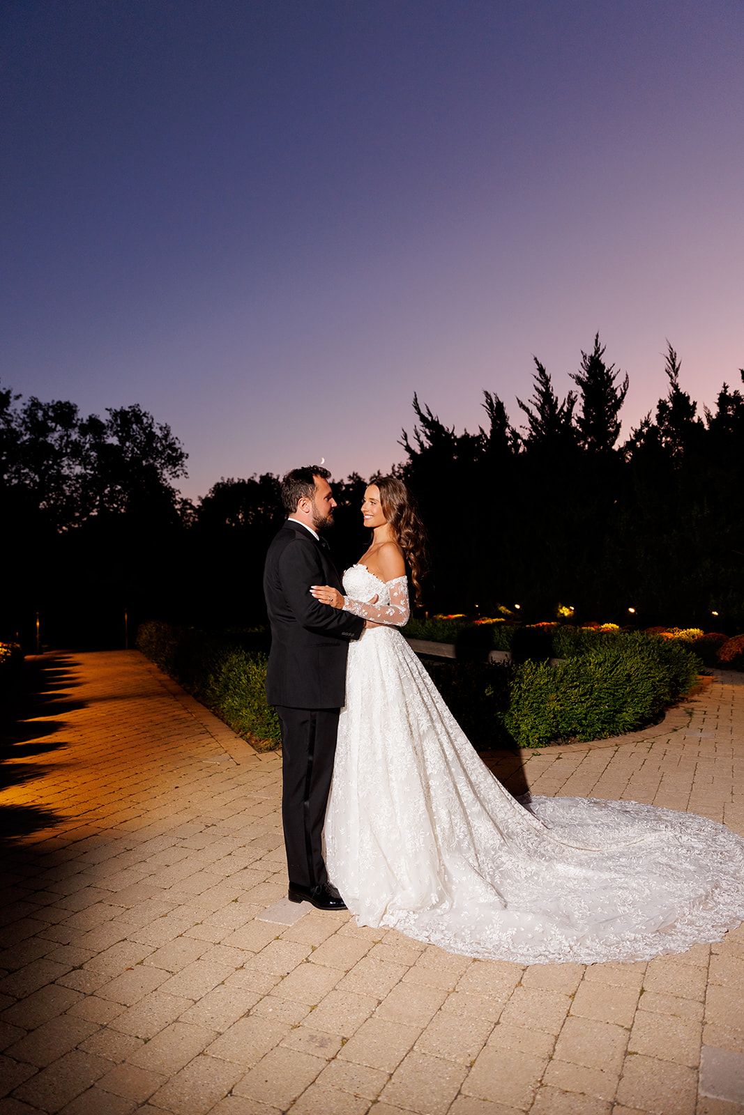Newlyweds share a romantic moment on a brick pathway at twilight with silhouetted trees behind them.