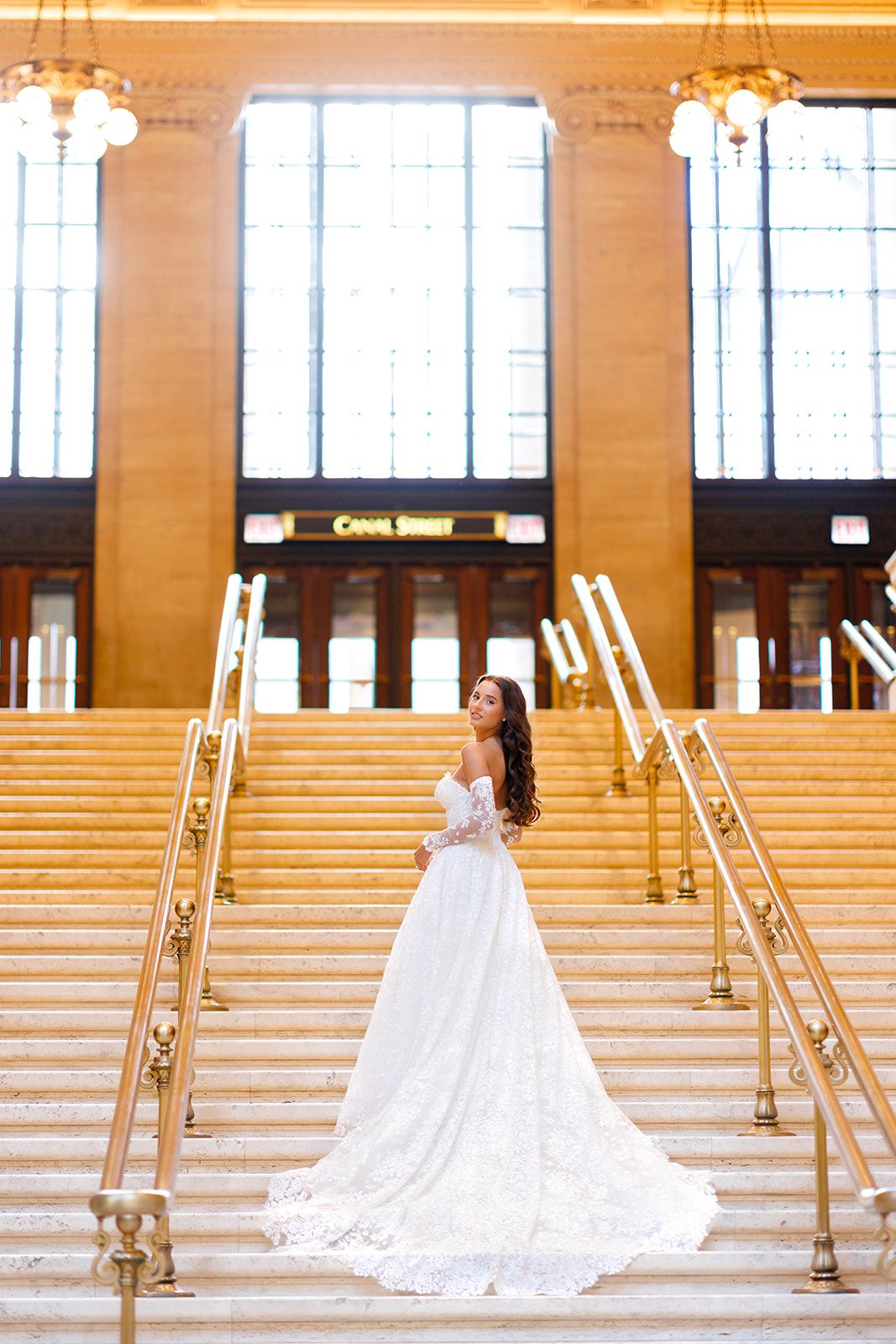 Bride in white lace gown with long train poses on grand staircase at Canal Street station.