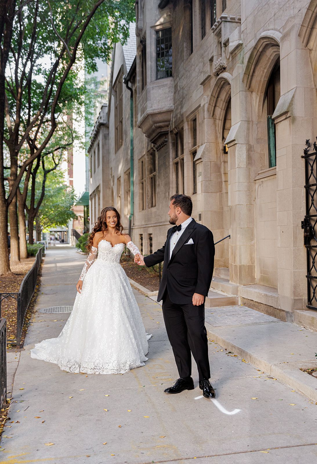 Bride in white lace gown and groom in tuxedo holding hands on urban sidewalk near stone building.