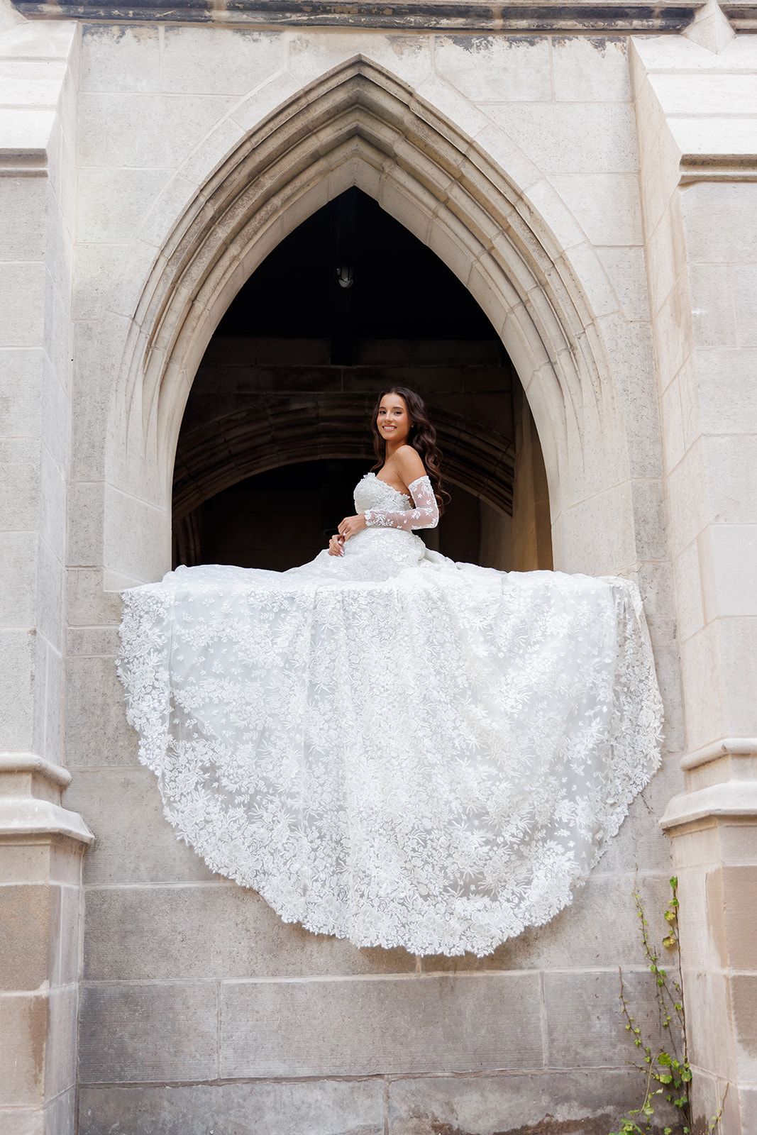 Bride in elegant white lace ballgown posing in Gothic stone archway, dress dramatically spread wide.
