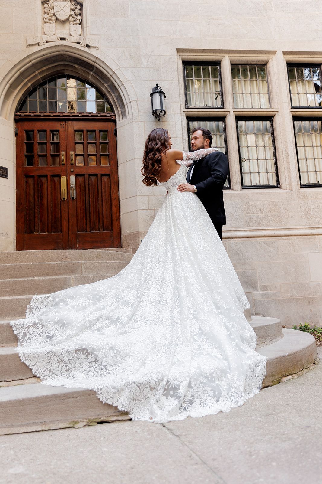 Bride in a stunning lace ball gown with a long train stands with groom on stone steps before ornate wooden doors.