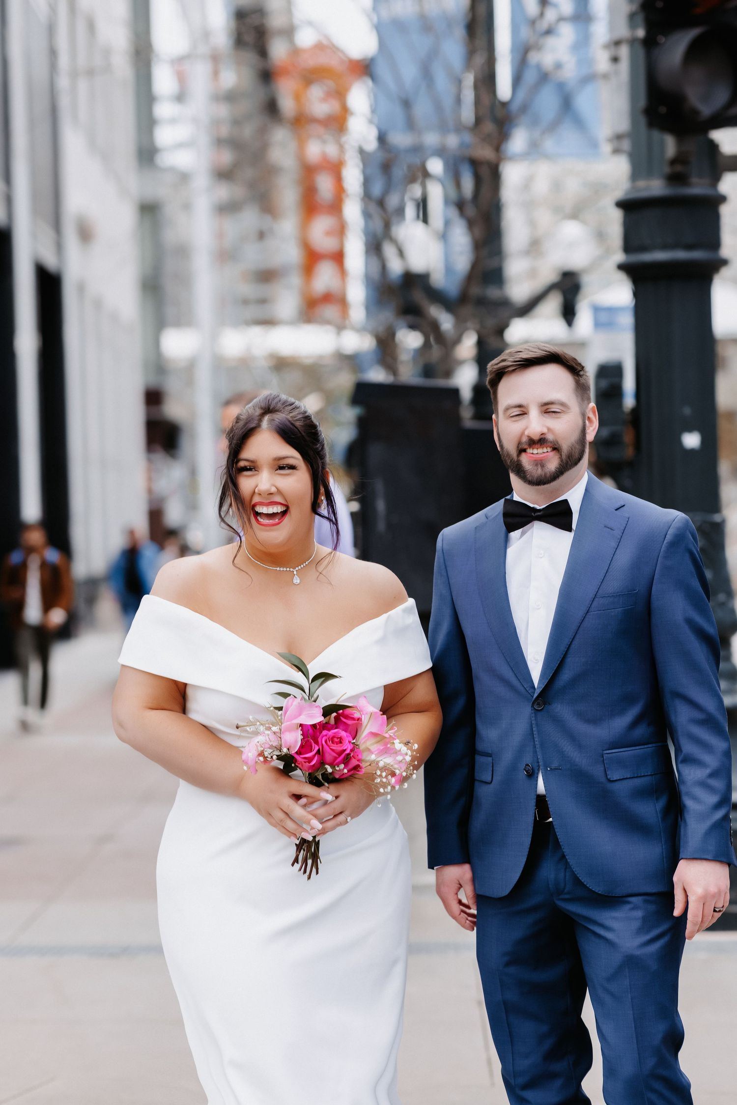 Joyful bride in off-shoulder white gown laughs beside bearded groom in navy suit on Chicago city street.