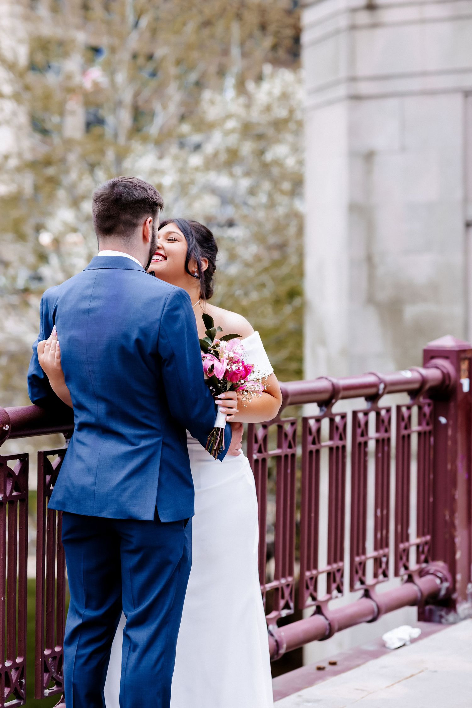 Bride in white off-shoulder gown laughs joyfully with groom in blue suit on ornate bridge with blooming trees.