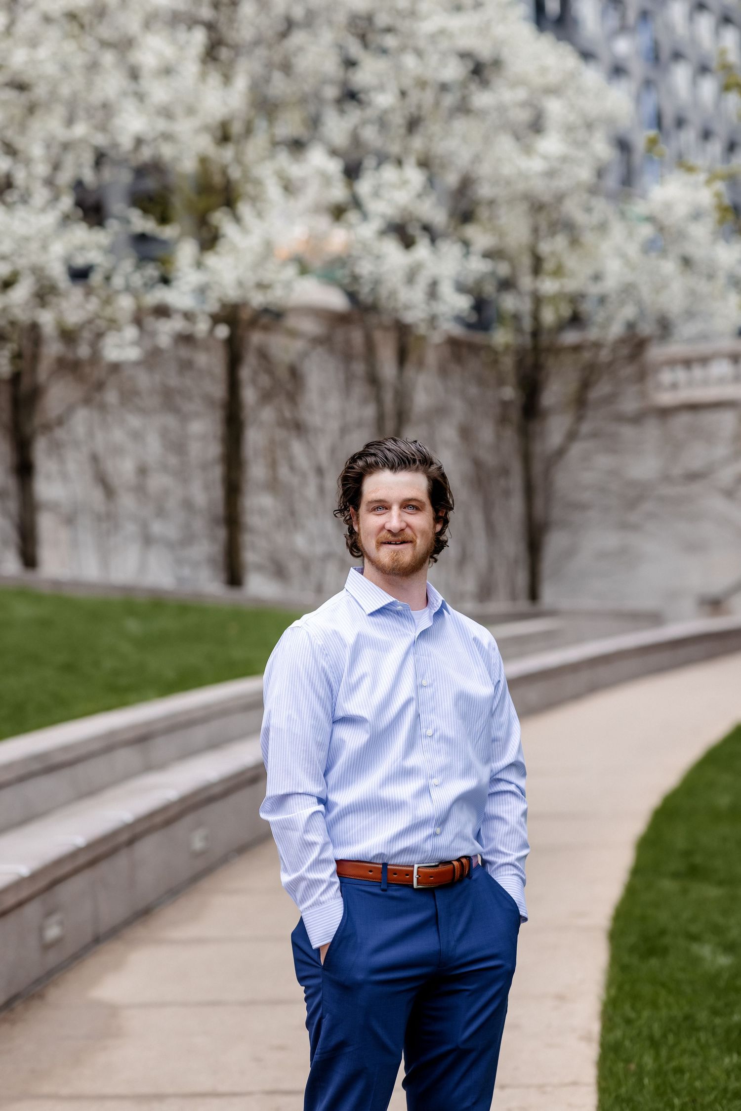 Smiling man in blue pants and light blue shirt stands on a path near white flowering trees.