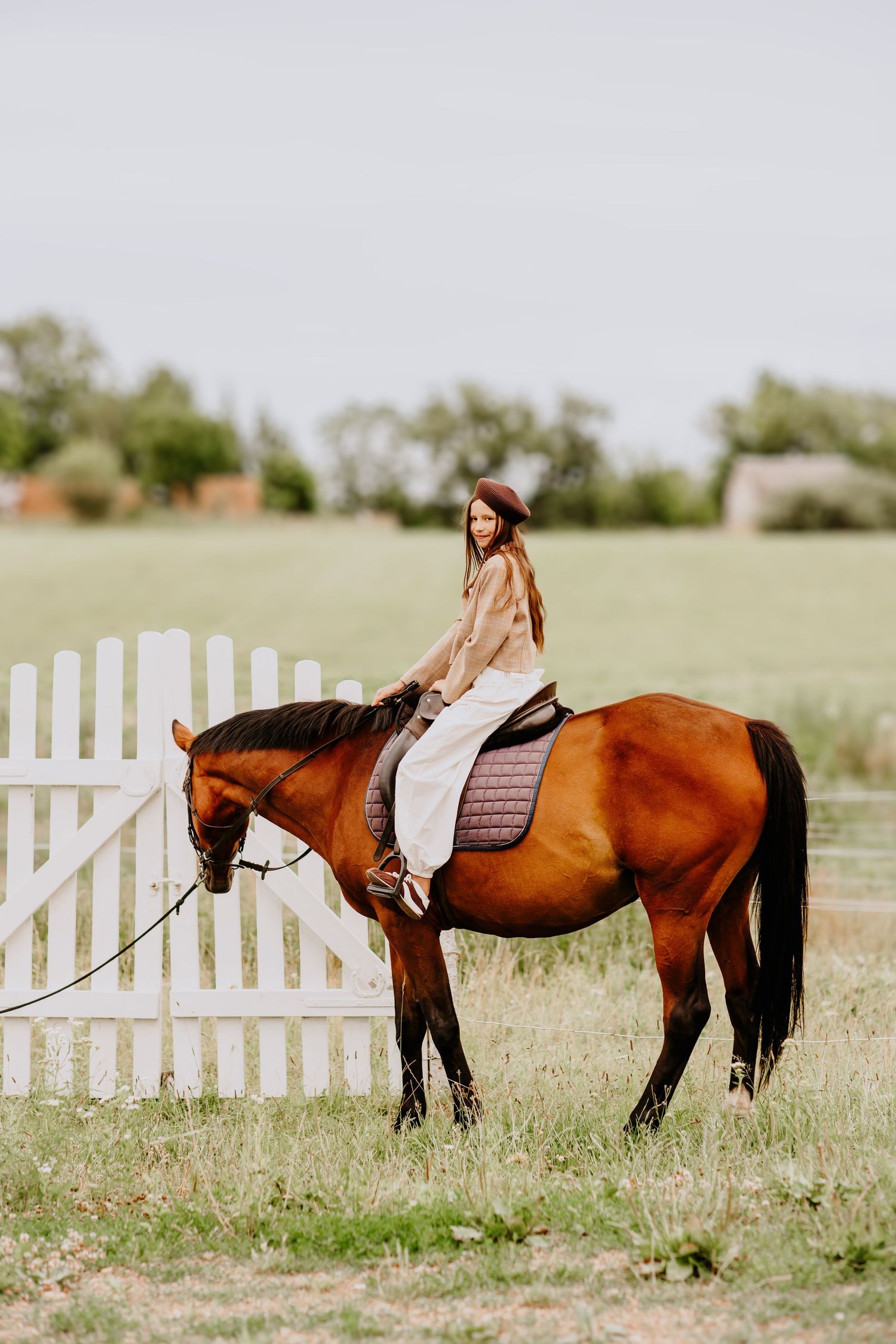 Young woman on a chestnut horse near a white picket fence on a farm.