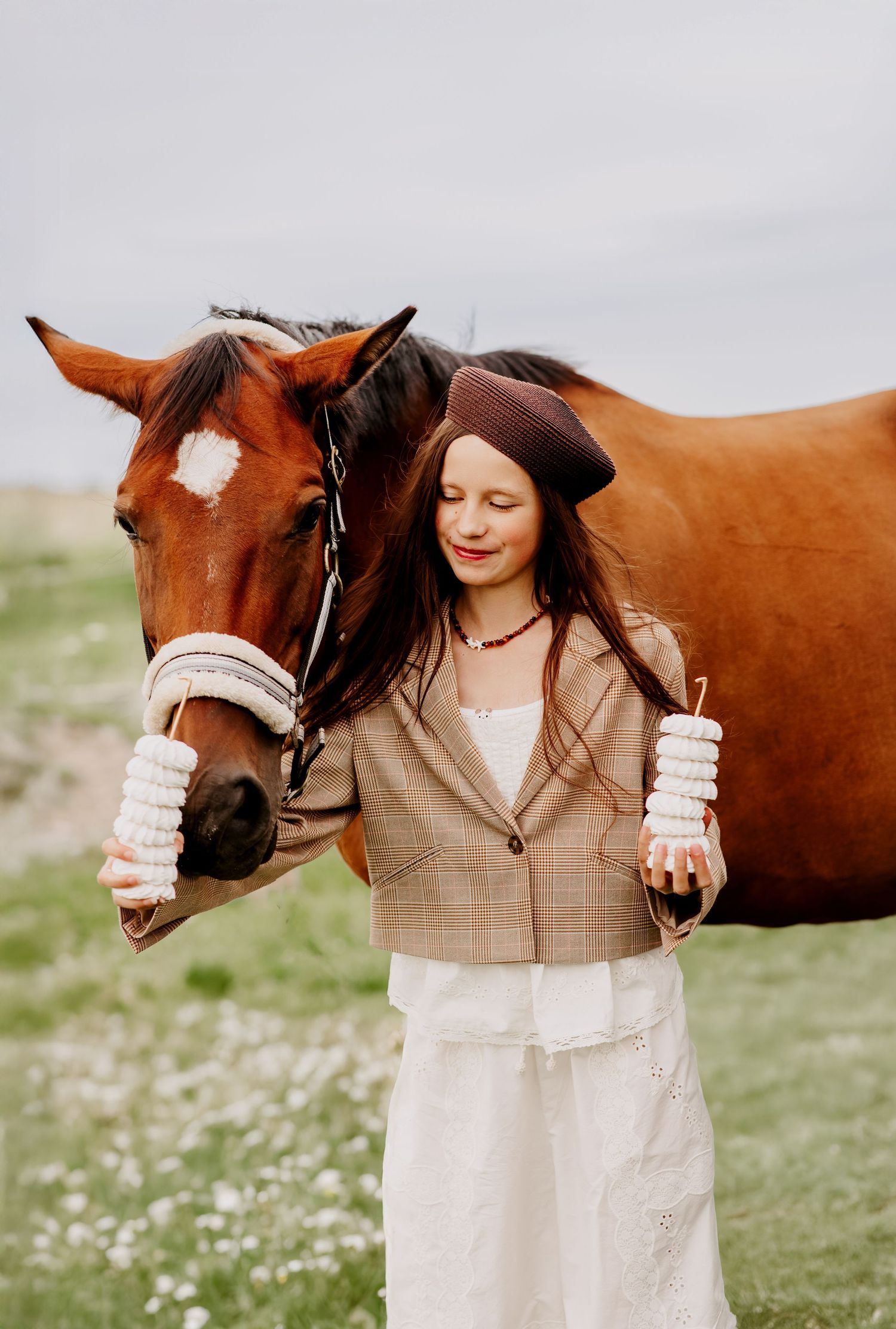 Young girl in brown beret and plaid blazer standing beside a chestnut horse in a green meadow with closed eyes.