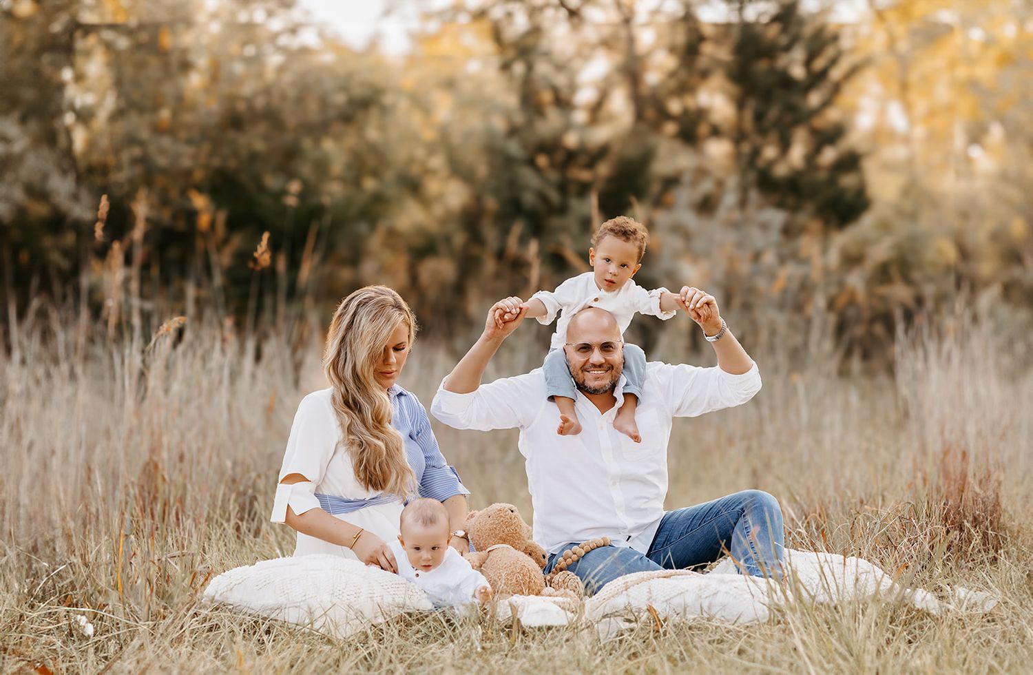 A happy family sits on a white blanket in tall grass, dad balancing a toddler on his shoulders with a baby nearby.