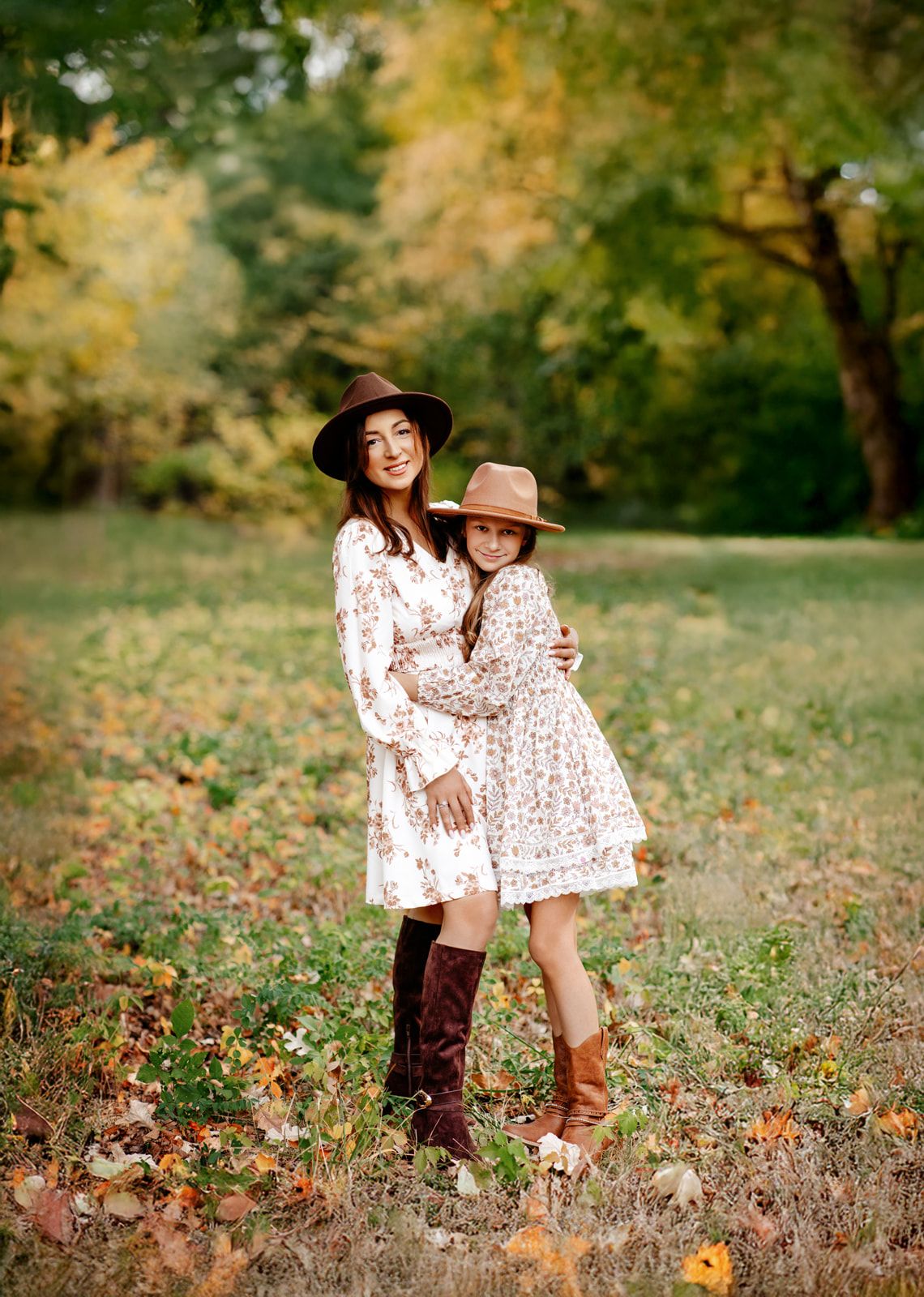 Mother and daughter in matching floral dresses and cowboy hats embrace in autumn park.