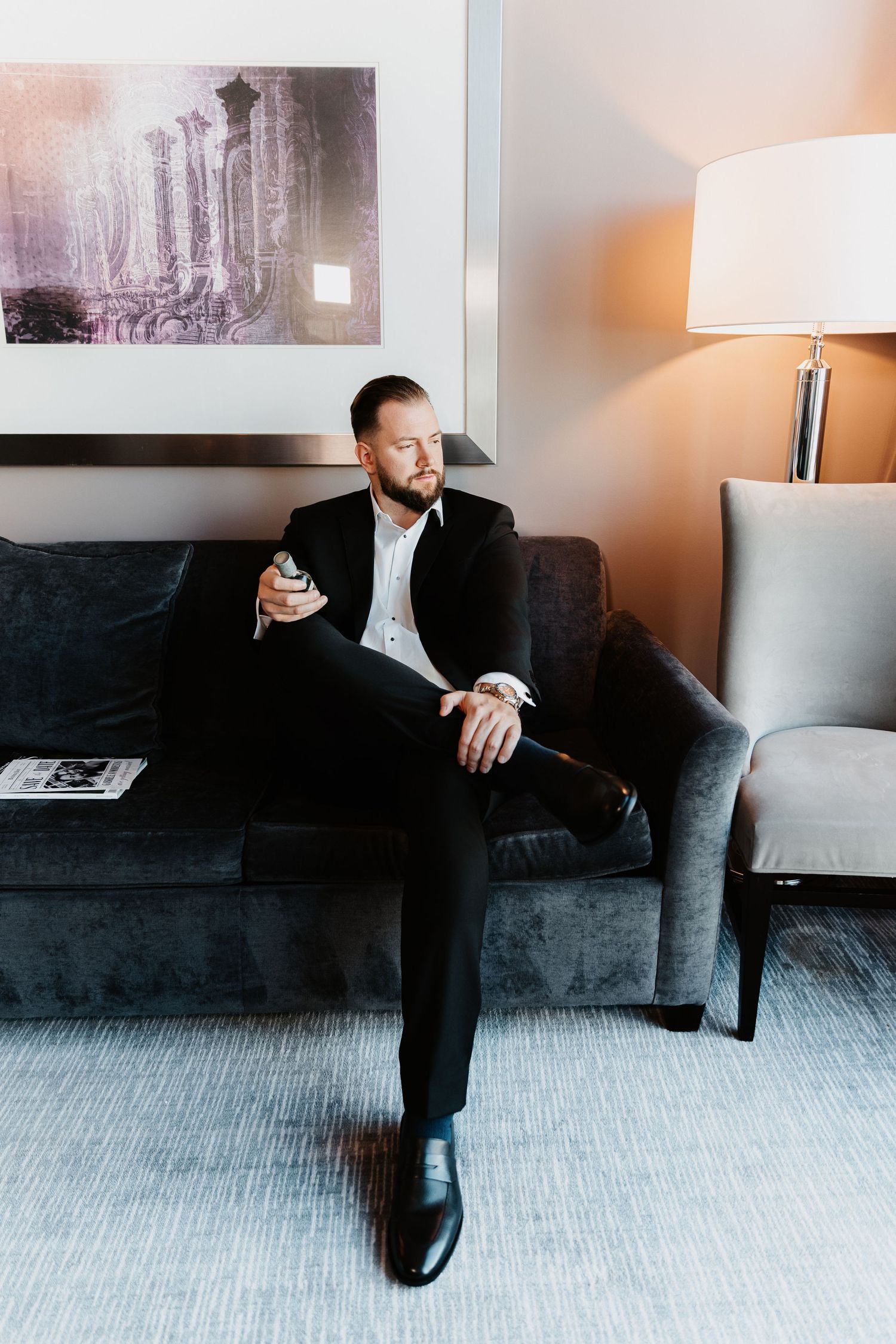 Groom in black suit checking watch while holding drink, seated on gray sofa in hotel room.