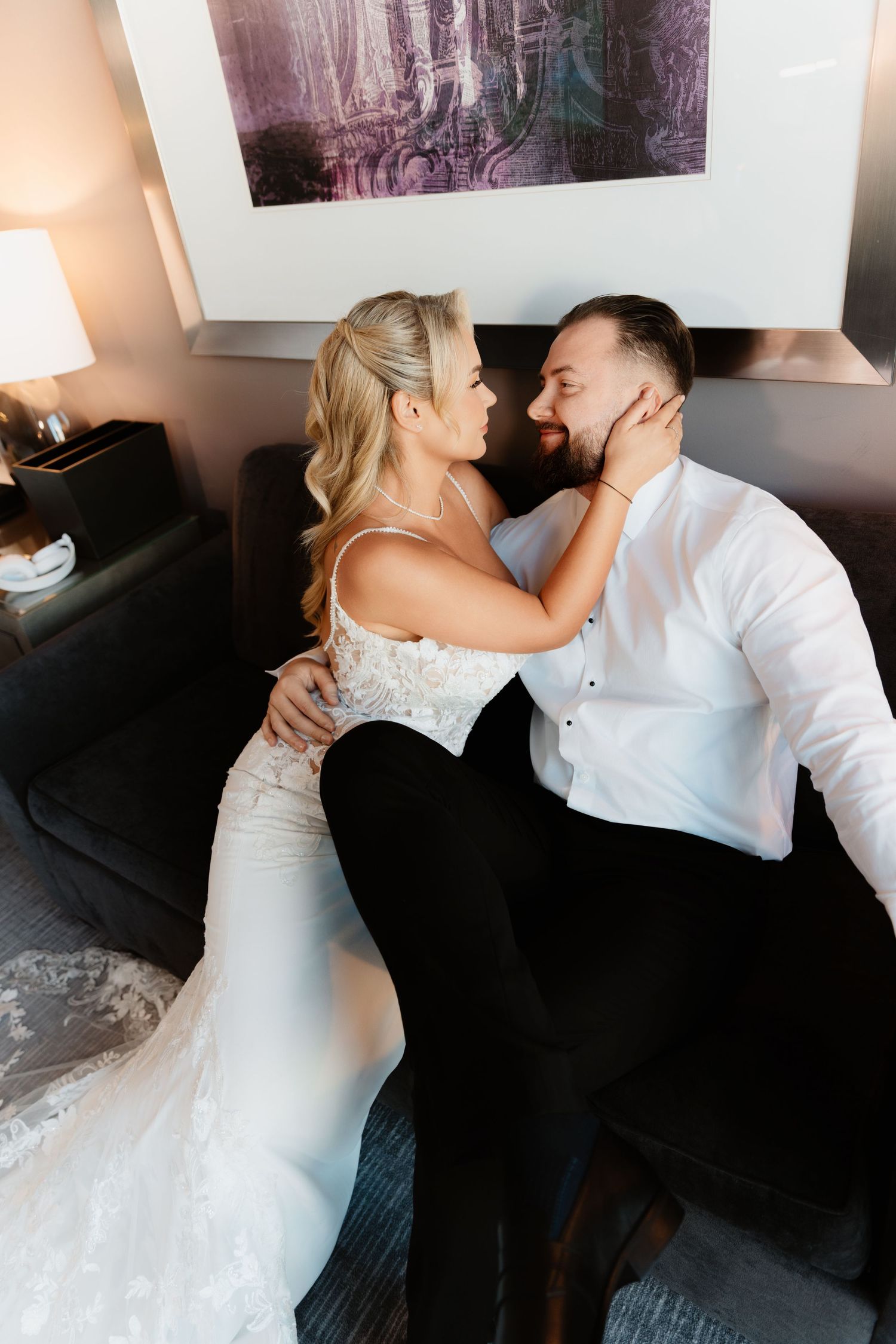 Bride in lace mermaid gown and groom share tender moment on sofa in warmly lit hotel suite.