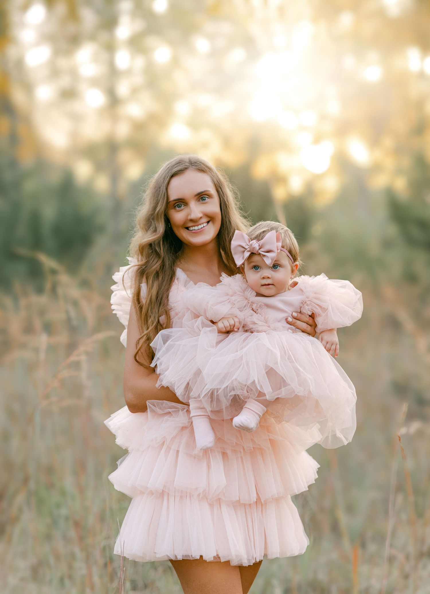 Smiling mother holding baby girl in matching blush pink tulle dresses in sunlit meadow.