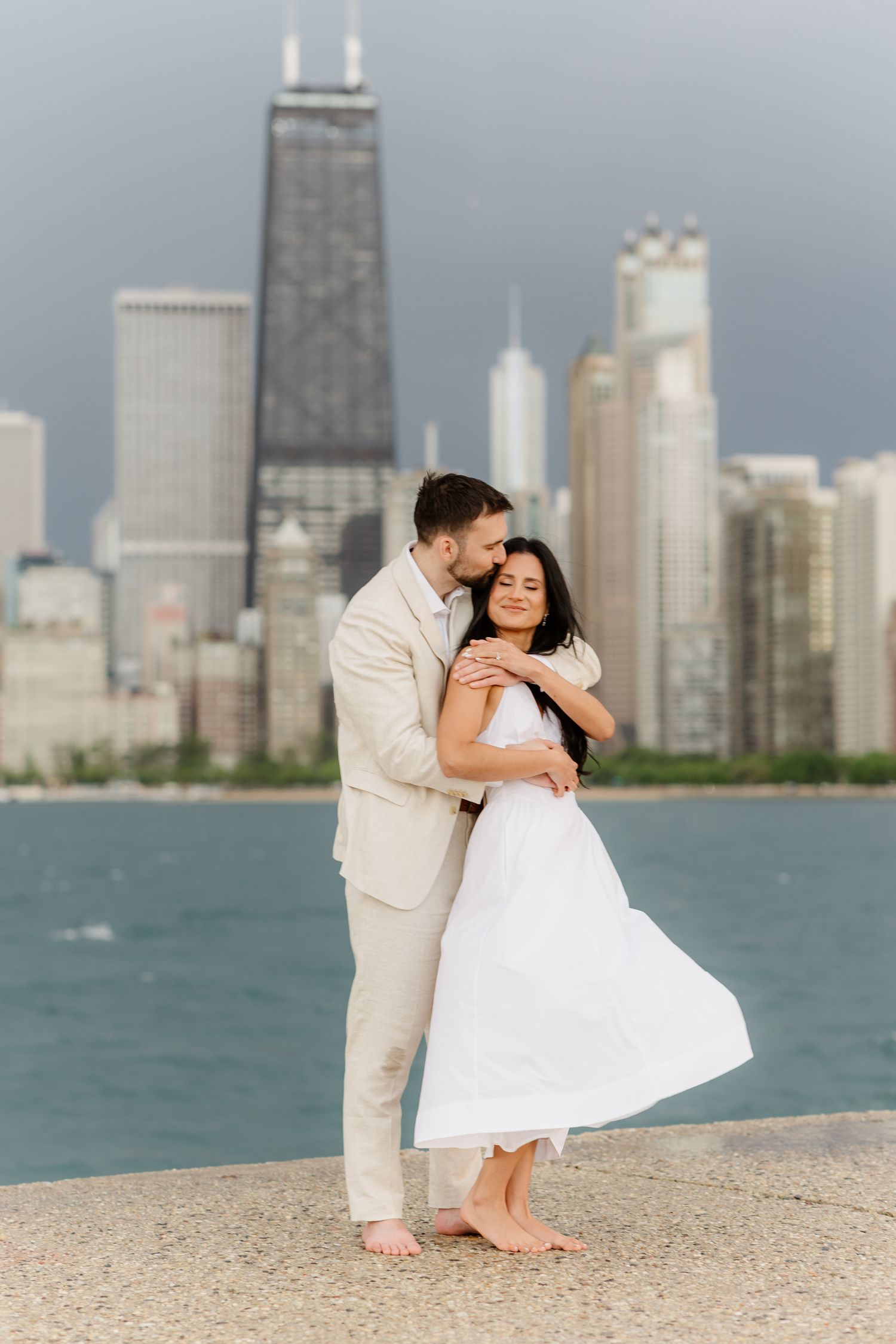 Couple sharing tender embrace on Chicago lakefront pier, woman in flowing white dress with city skyline background.