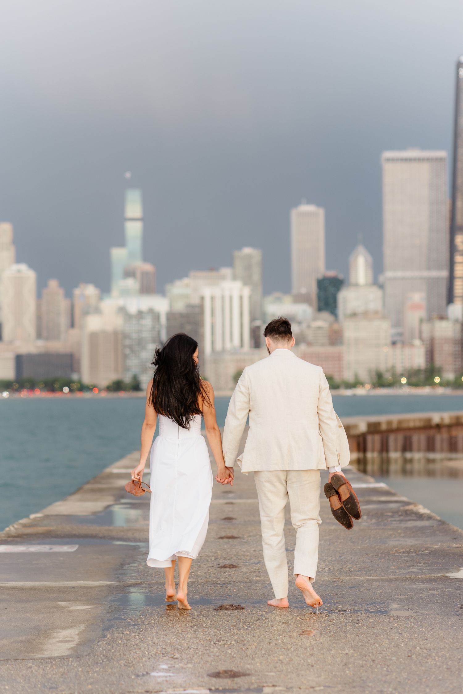 Couple walks hand-in-hand away from camera barefoot on wet pier with Chicago skyline at dusk.