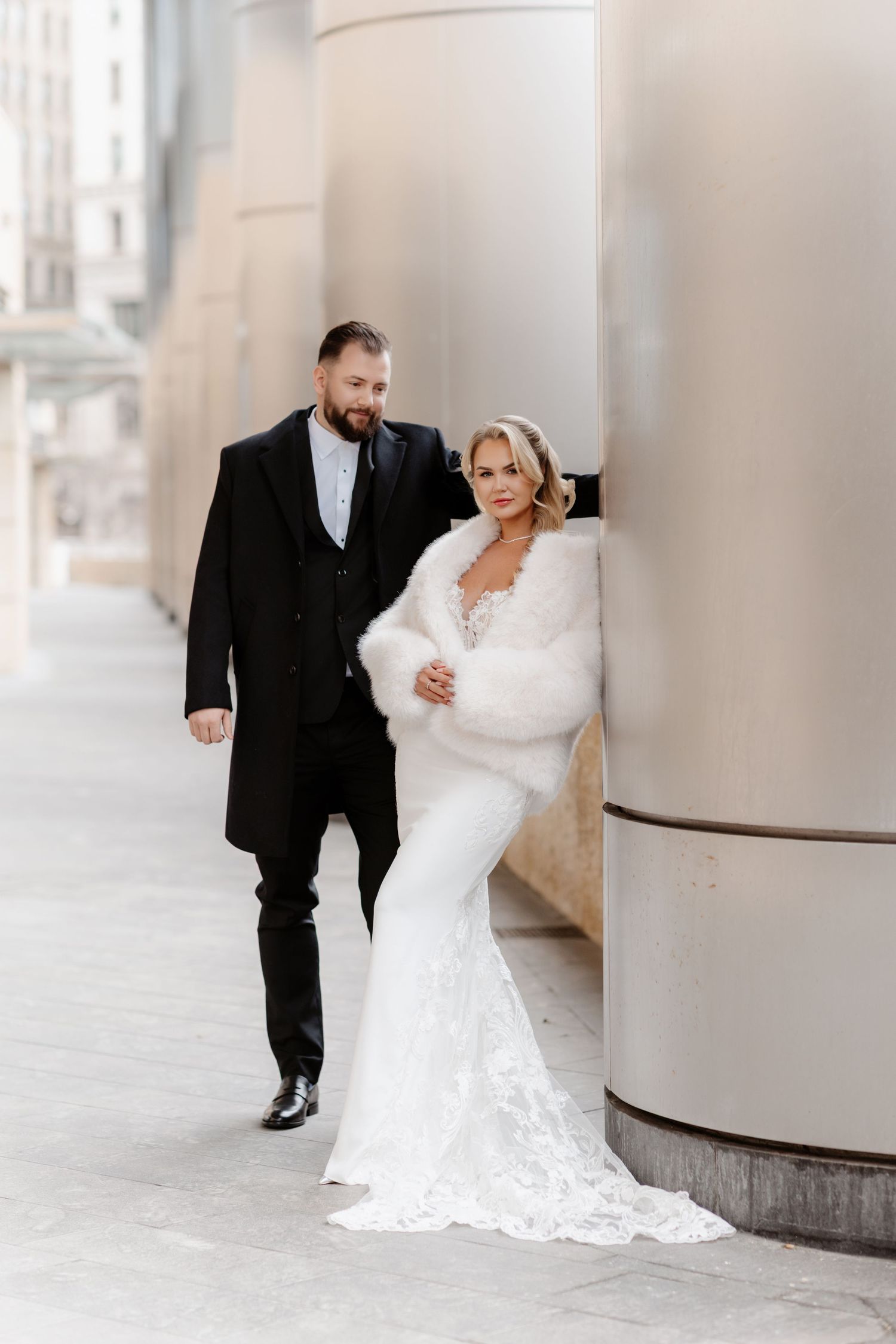 Bride in lace mermaid gown with white fur stole and groom in black suit by city building columns.