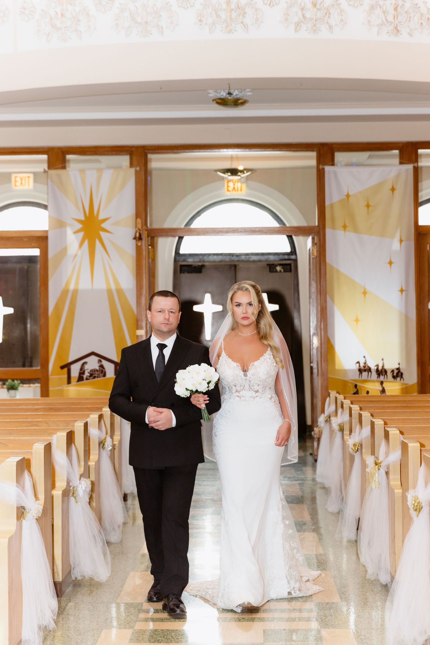 Bride in white gown walks down church aisle with groom in black suit, flanked by pews with white tulle decorations.