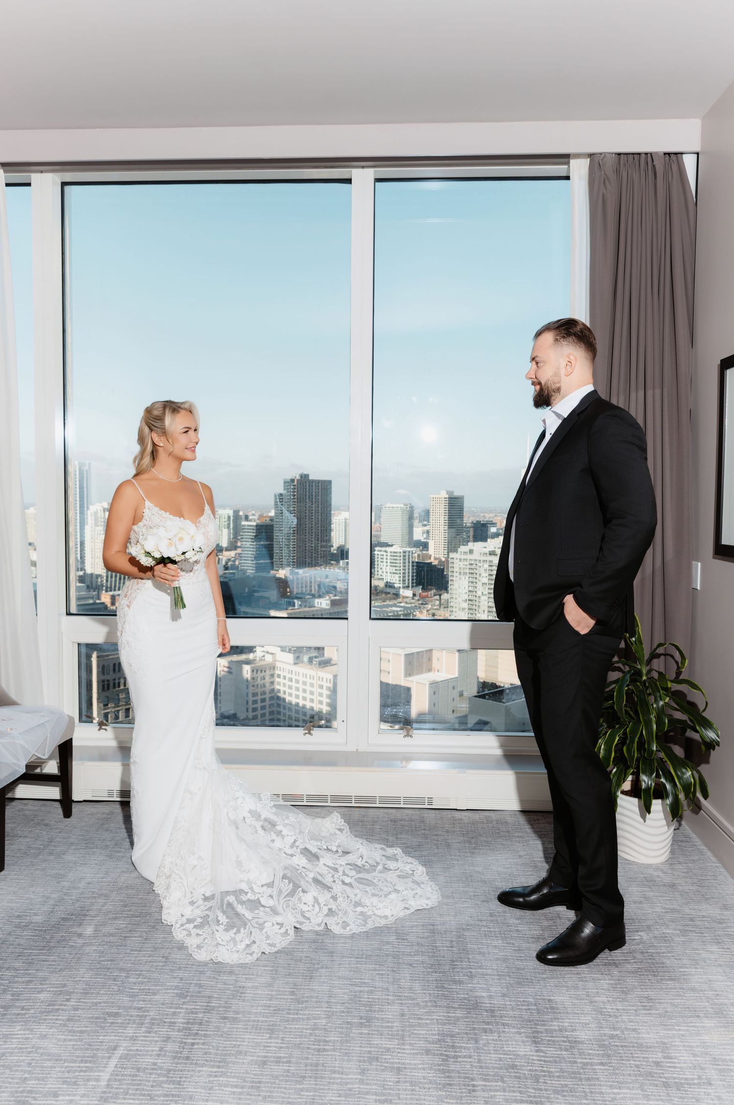 Bride in lace wedding gown smiles at groom in black tuxedo during first look in hotel suite.