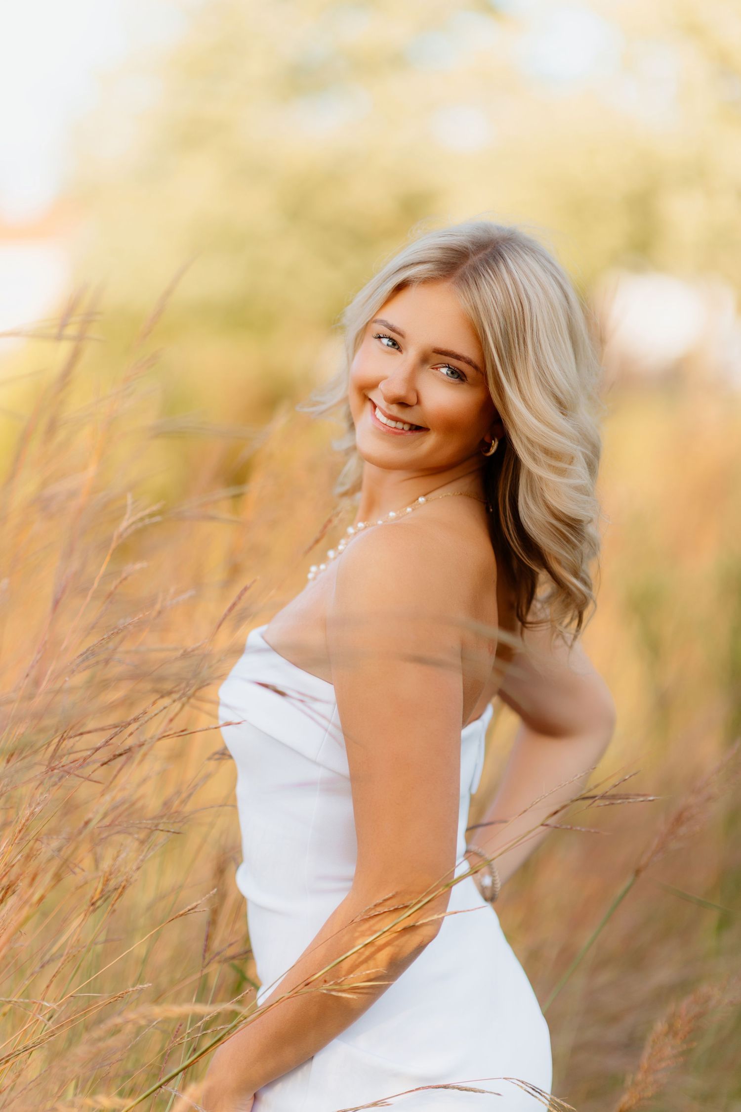 Blonde woman in white strapless dress smiling over shoulder in golden tall grass field.