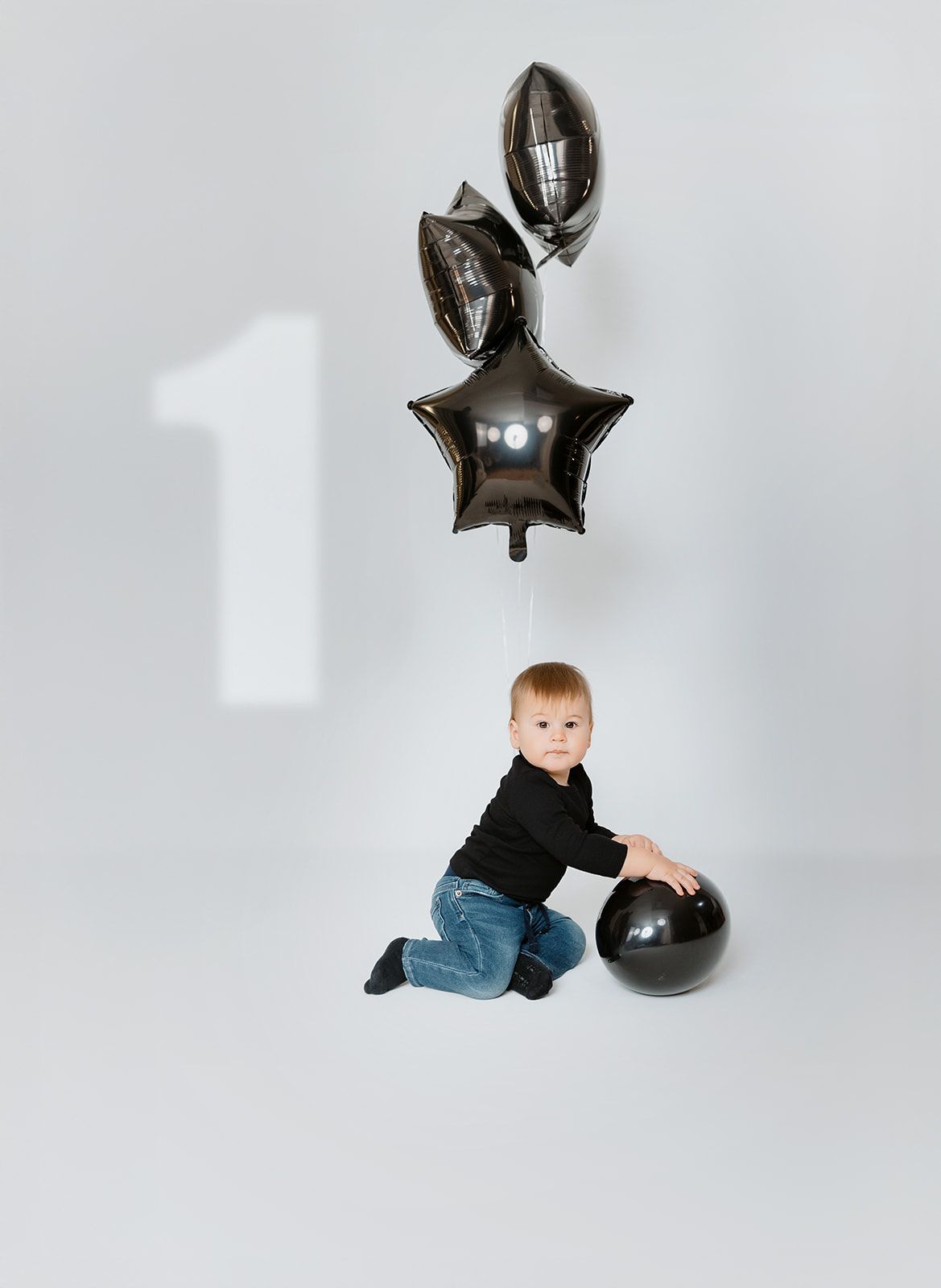 A toddler in black sweater and jeans sits with black balloons and a large black ball, celebrating his first birthday.