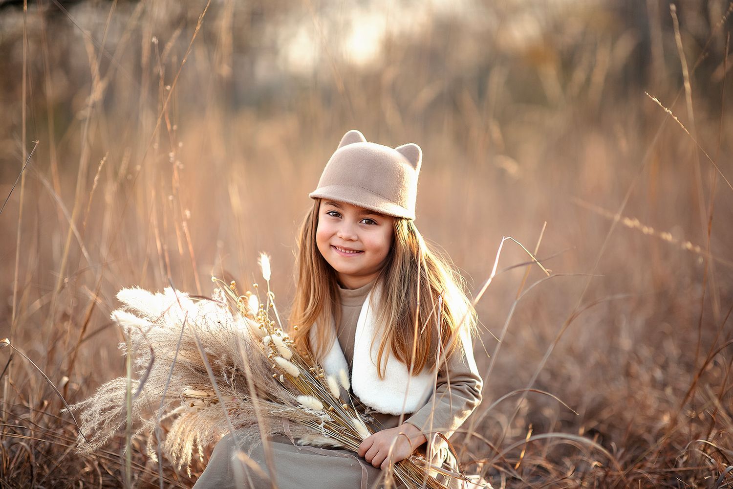A smiling girl in a bear-ear hat holds dried wheat in a sunlit autumn field.