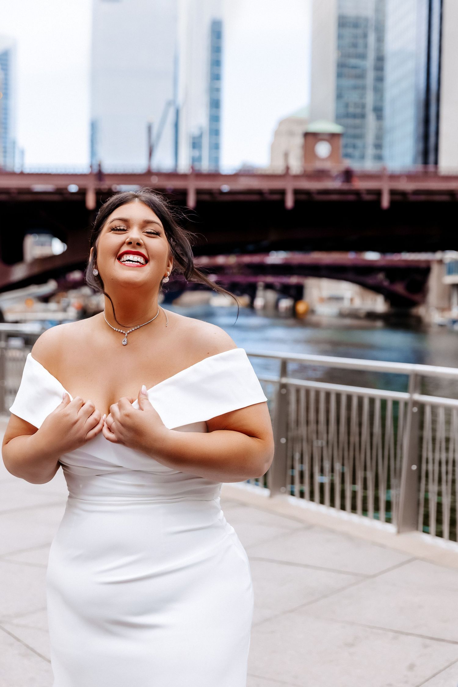 Bride in off-shoulder white gown laughing joyfully on Chicago Riverwalk with city skyline and bridge backdrop.