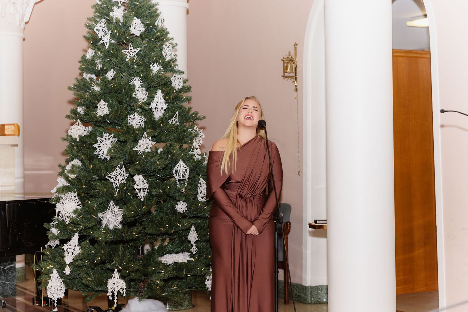 Woman in brown dress poses beside decorated Christmas tree with white snowflake ornaments indoors.