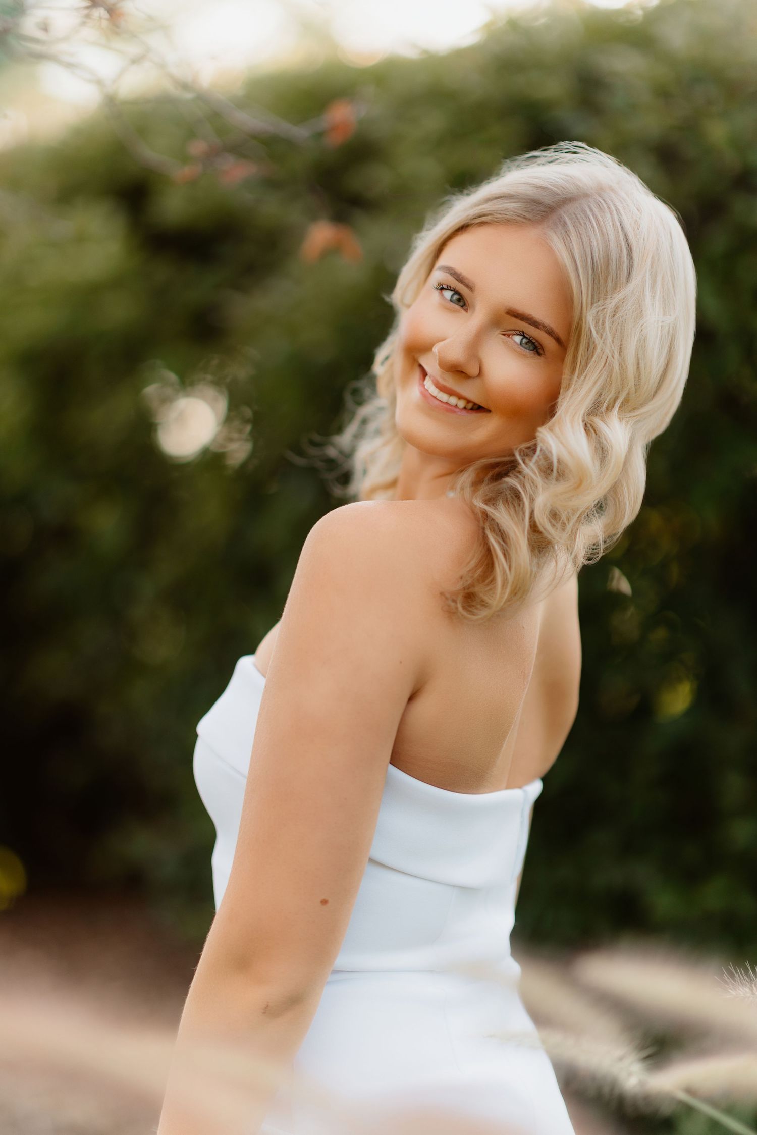 Blonde woman in white strapless dress smiling with green foliage background outdoors.